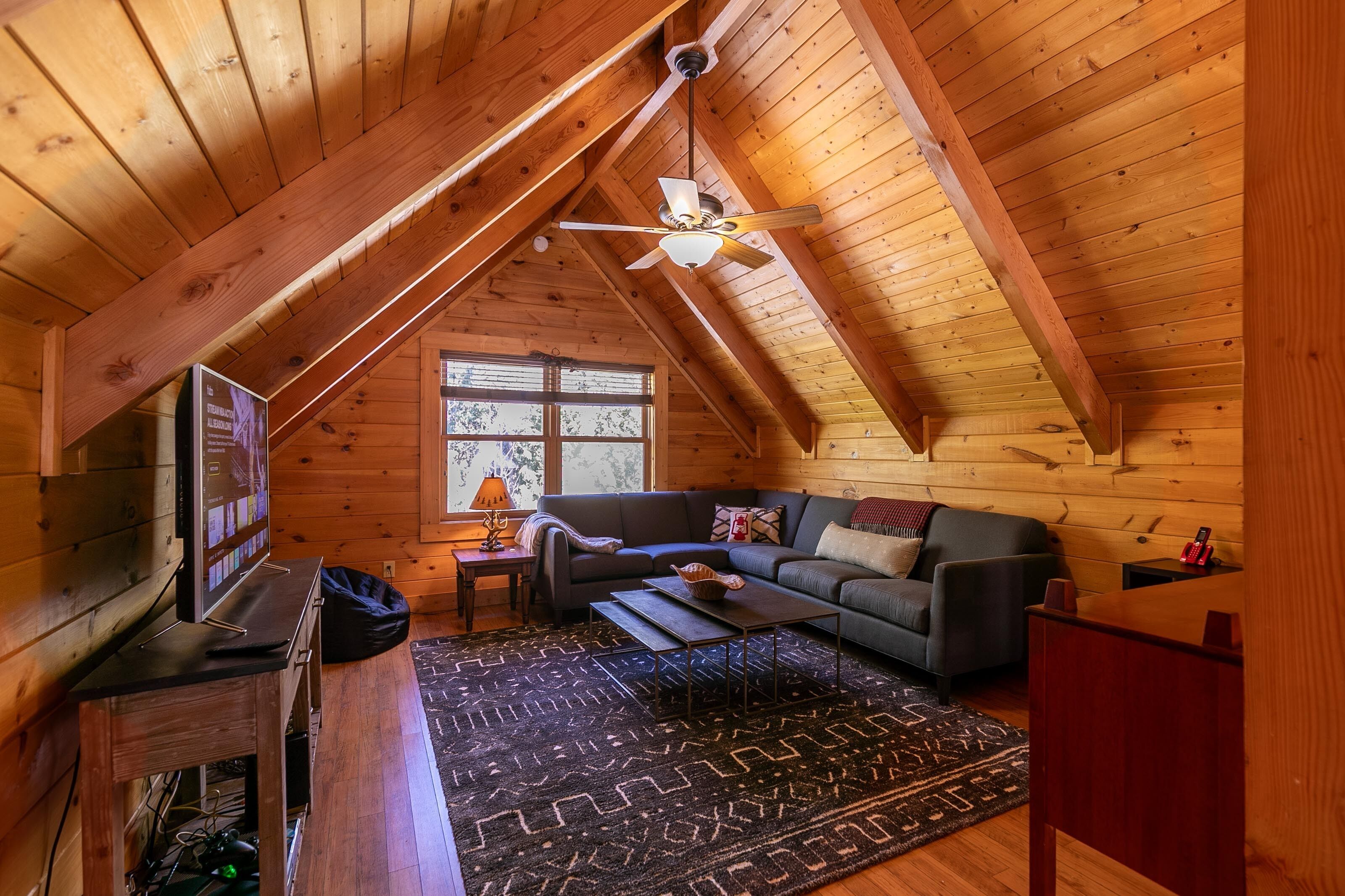 A cozy attic room with wooden walls and a sloped ceiling. It features a dark-colored sofa, a small table, and a television on a wooden stand.