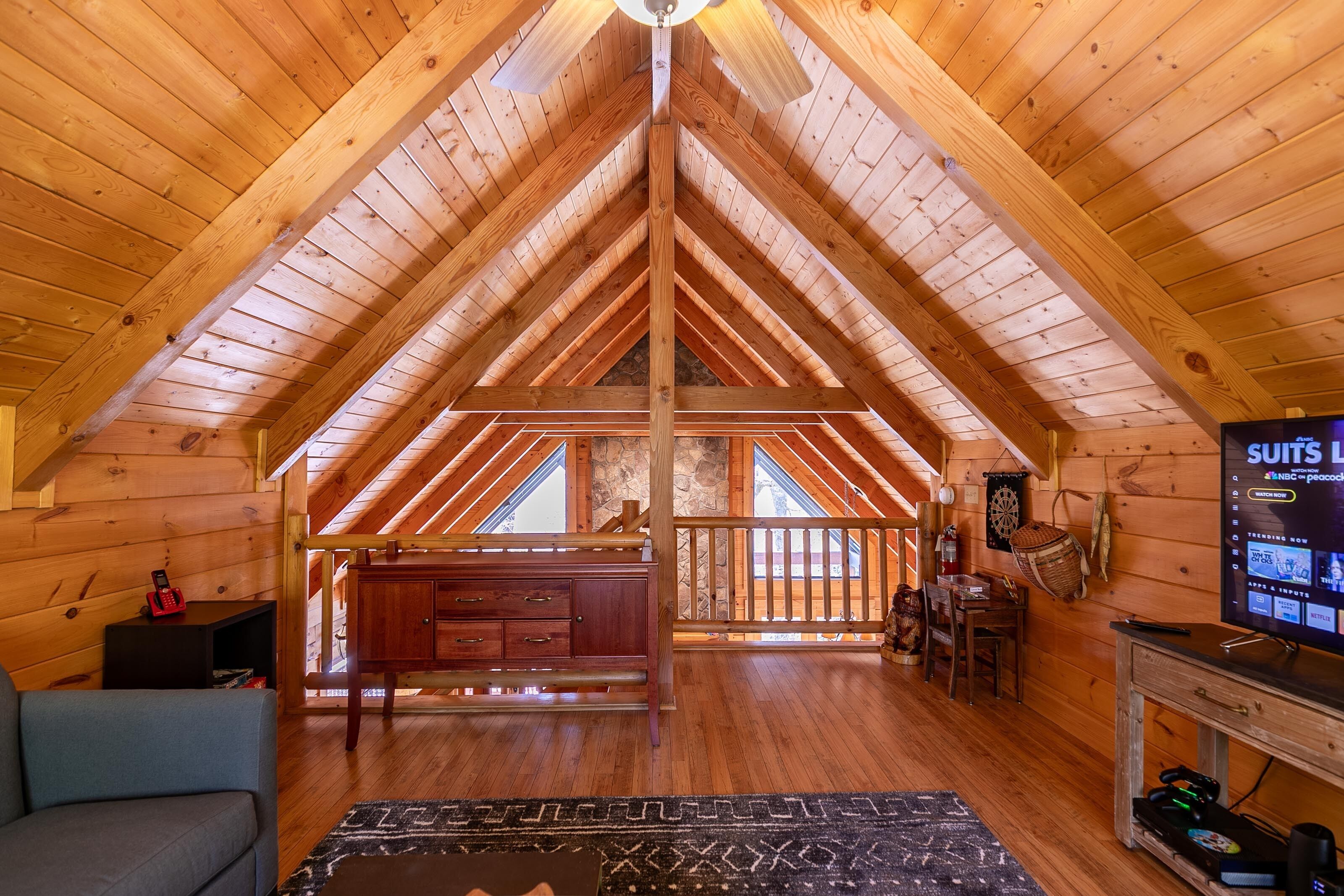 A spacious attic room with wooden beams, a wooden ceiling, a television, and a view through the windows.