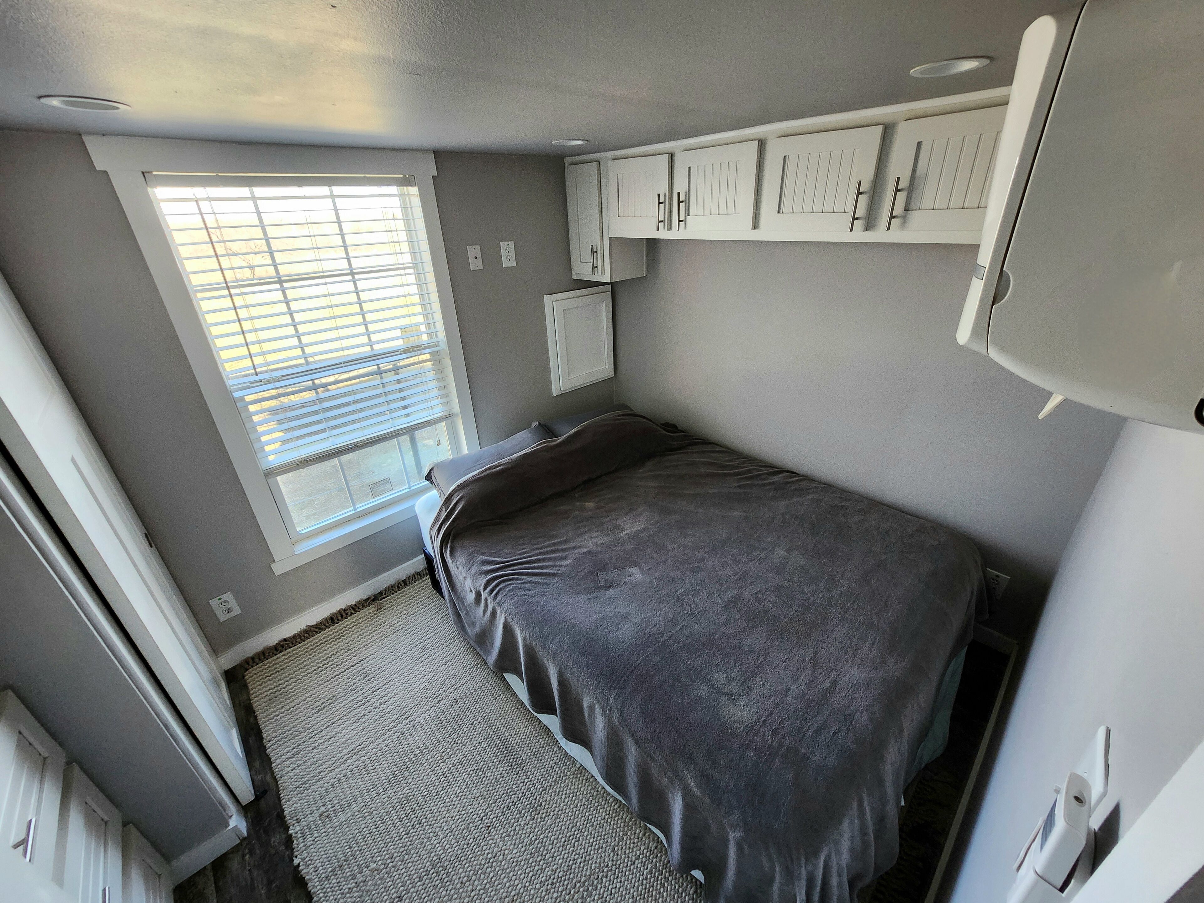 A compact bedroom with a bed, a window with blinds, and built-in cabinets.
