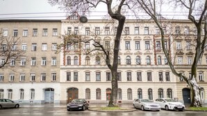 Ein mehrstöckiges Gebäude im klassischen Stil, mit einem zentralen Eingang, einer großen Tür und zahlreichen Fenstern in jeder Etage.