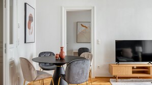 A modern living room with a round black table, grey upholstered chairs, a flat-screen TV, and a wooden TV stand.