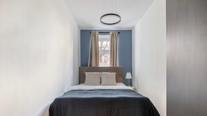 Een smalle, goed verlichte slaapkamer met een bed dat gecentreerd is onder een ronde lamp, een raam met gordijnen en een nachtkastje met een lamp.