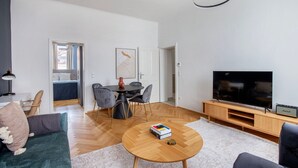 Een moderne woonkamer met een ronde houten salontafel, een flatscreen televisie op een houten standaard, een eethoek met een ronde tafel en stoelen, en een gezellige zithoek met een bank en fauteuils.