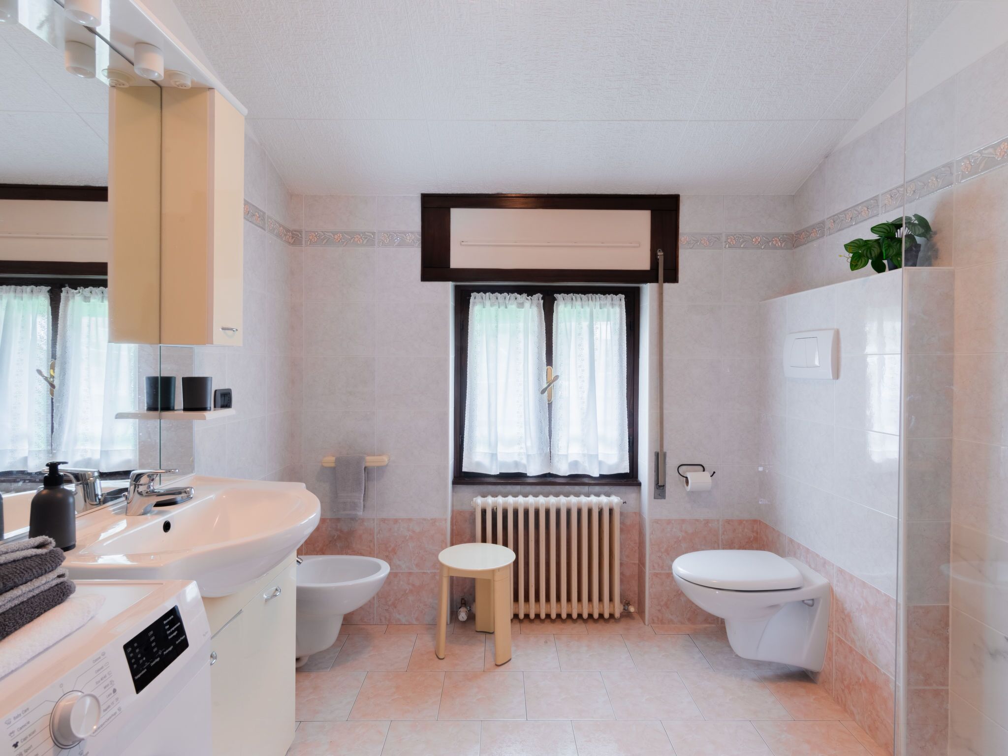 A modern bathroom with a sink, toilet, and bidet. There is a window with curtains, a radiator, and a small stool.