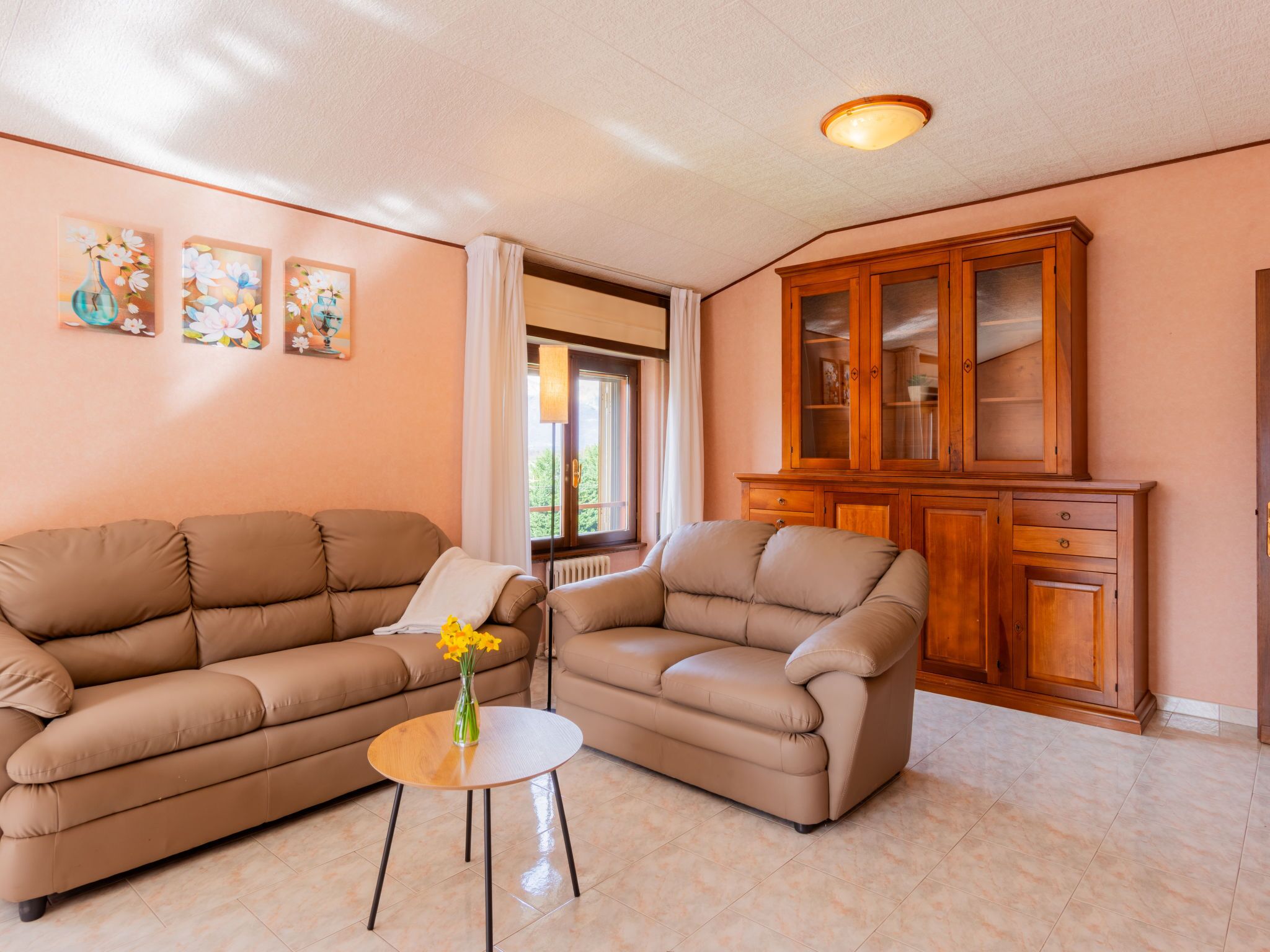 A living room with a beige sofa, a small round table with a vase of flowers, and a wooden cabinet.