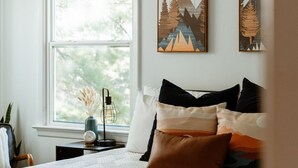 Ein Schlafzimmer mit großem Fenster, einem Bett mit vielen Kissen, einem Nachttisch mit Lampe und einer Uhr sowie zwei an der Wand hängenden Bildern von Berglandschaften.