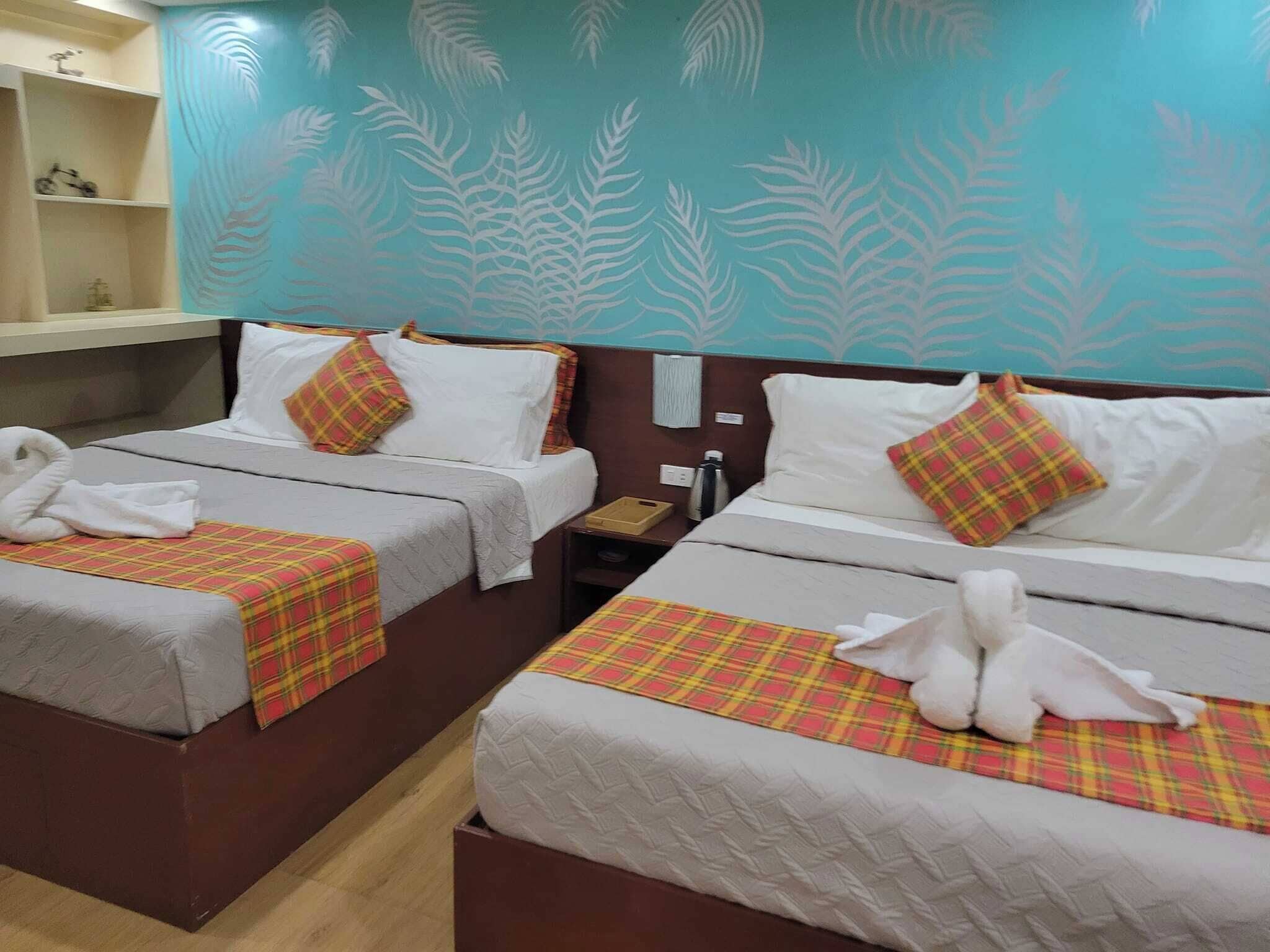 Habitación de hotel con dos camas, cada una con manta y almohadas a cuadros, una mesita con una botella, y una pared con diseño de hojas.