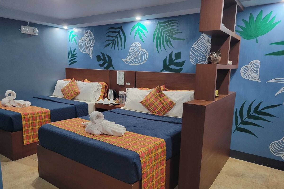 A hotel room with two beds, each with a blue bedspread and a plaid blanket. The room has a blue wall with leaf patterns and a wooden headboard.