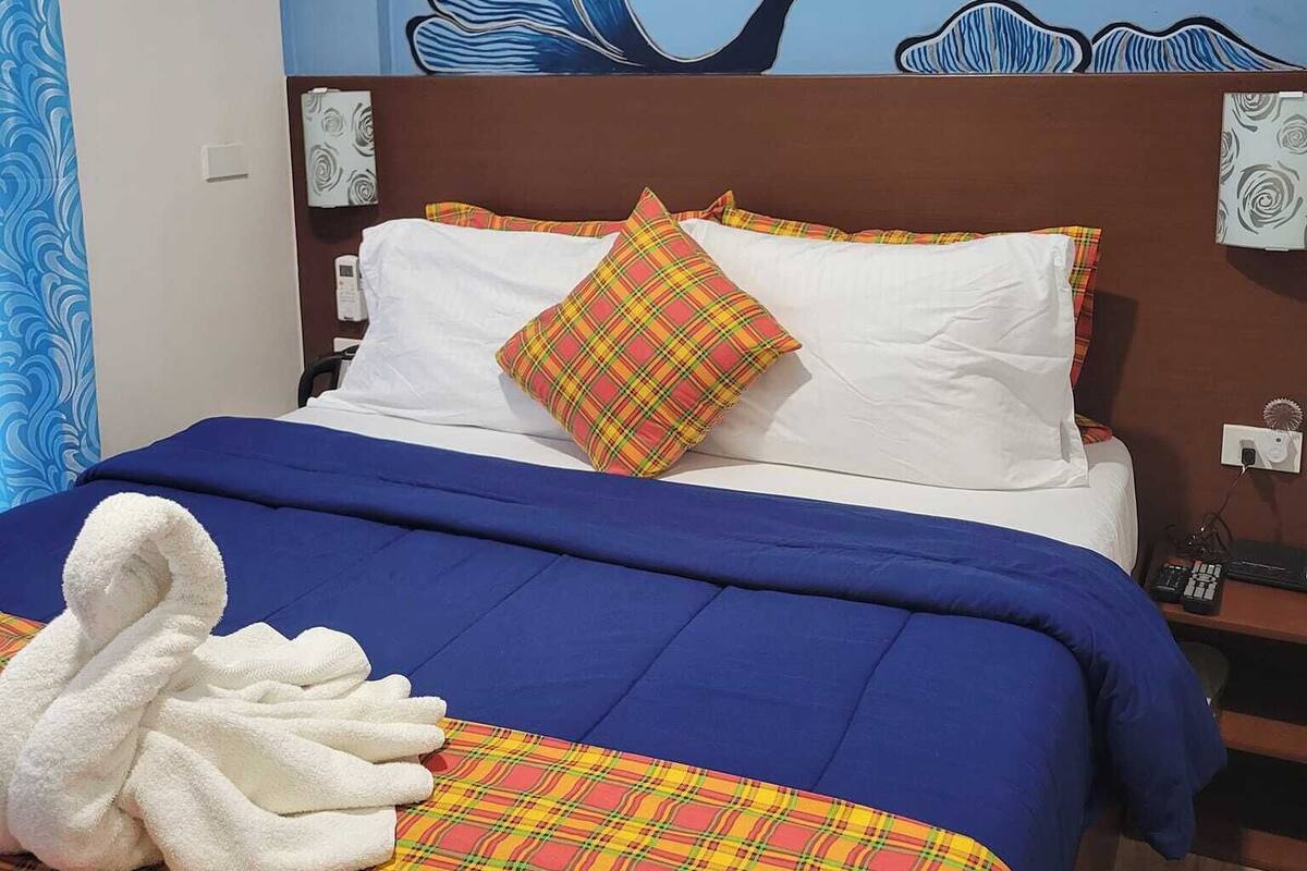 A hotel room with a bed, a blue and white wall mural, a wooden headboard, and a bedside table with a phone and a radio.