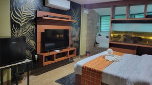 A hotel room with a flat-screen TV, a bed with a plaid blanket, a small table with a TV, and a window with a view of a pool. - Costa Madrangca (San Jose de Buenavista)
