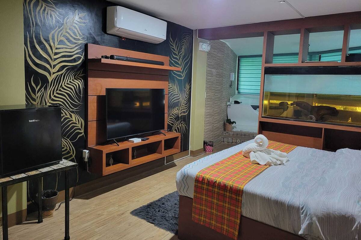 A hotel room with a flat-screen TV, a bed with a plaid blanket, a small table with a TV, and a window with a view of a pool.