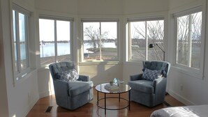 Una habitación con dos sillones, una mesita redonda y ventanas amplias con vistas al agua y árboles.