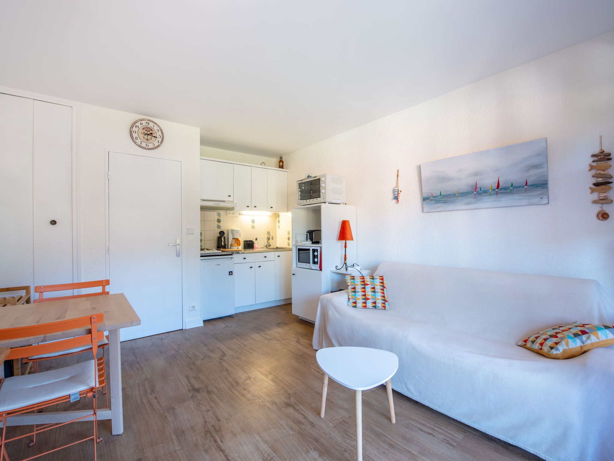 A compact living space with a kitchenette, a dining area with a table and chairs, a sofa with a patterned pillow, and a painting of sailboats on the wall.