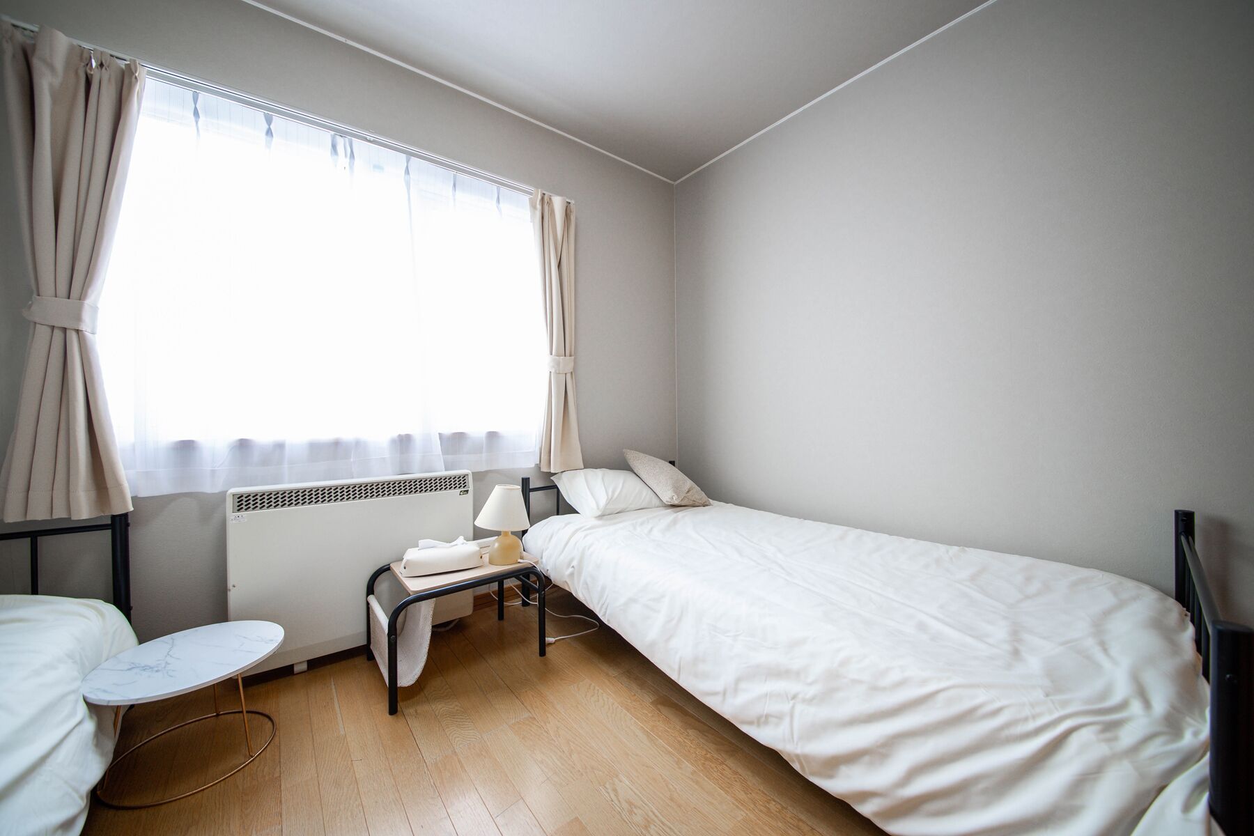 A small, clean room with a single bed, a bedside table, a chair, and a window with curtains.