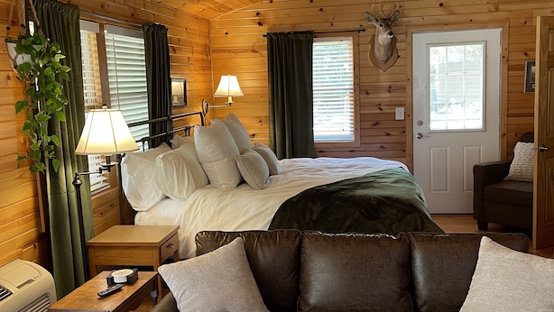 A cozy cabin interior with a wooden ceiling, a bed with white bedding, a brown leather sofa, a lamp, and a mounted deer head on the wall. - Cozy cabin in delightful Wisconsin Dells- Cabin A (Wisconsin Dells)