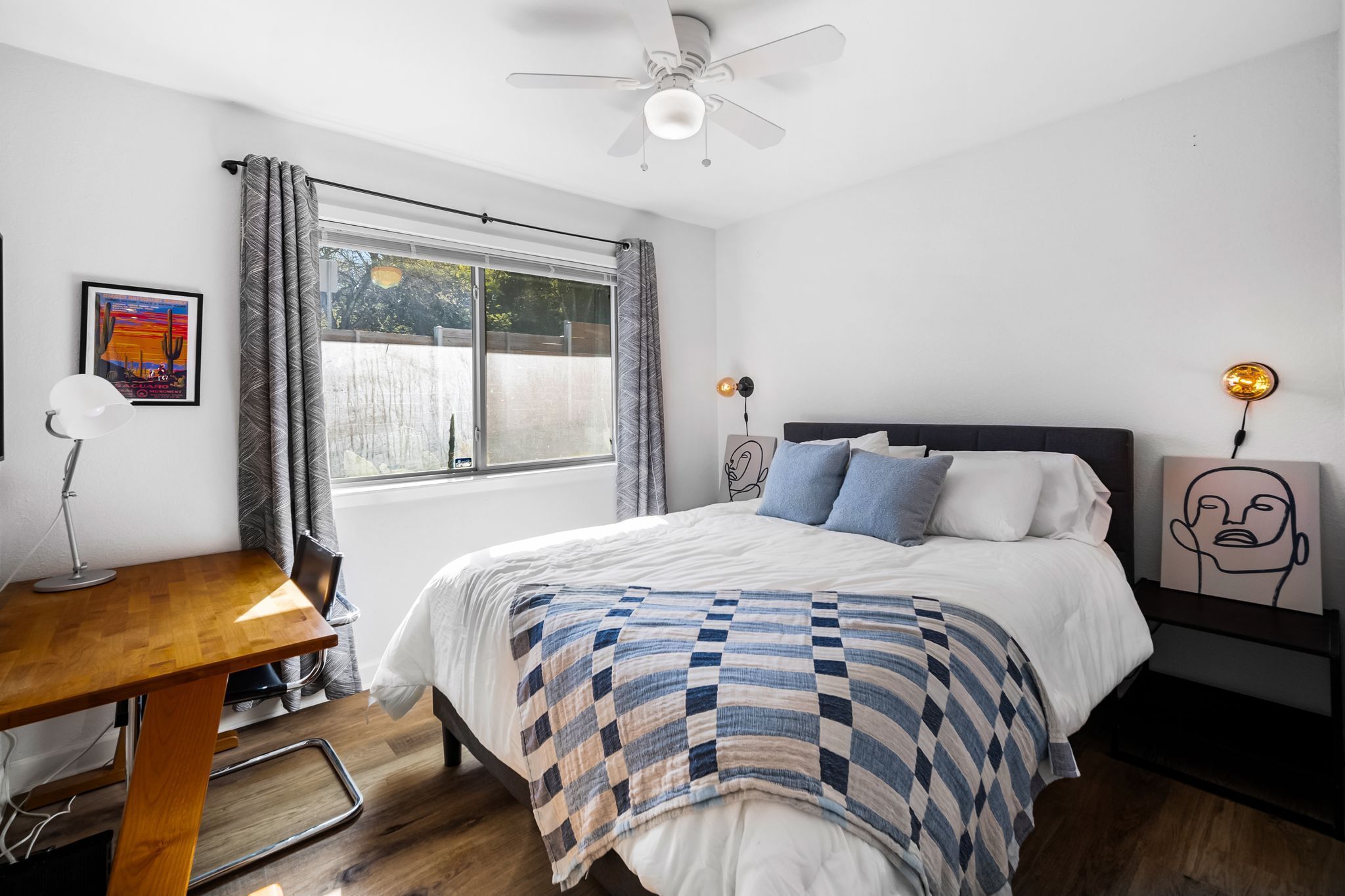 A bedroom with a bed, a desk, a chair, a ceiling fan, a window with curtains, and artwork on the walls.