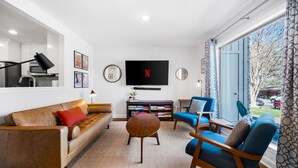 Un salon moderne avec un canapé en cuir, une table basse ronde et des fauteuils bleus. Un téléviseur à écran plat est fixé au mur, et il y a une bibliothèque remplie de livres et d’objets décoratifs.