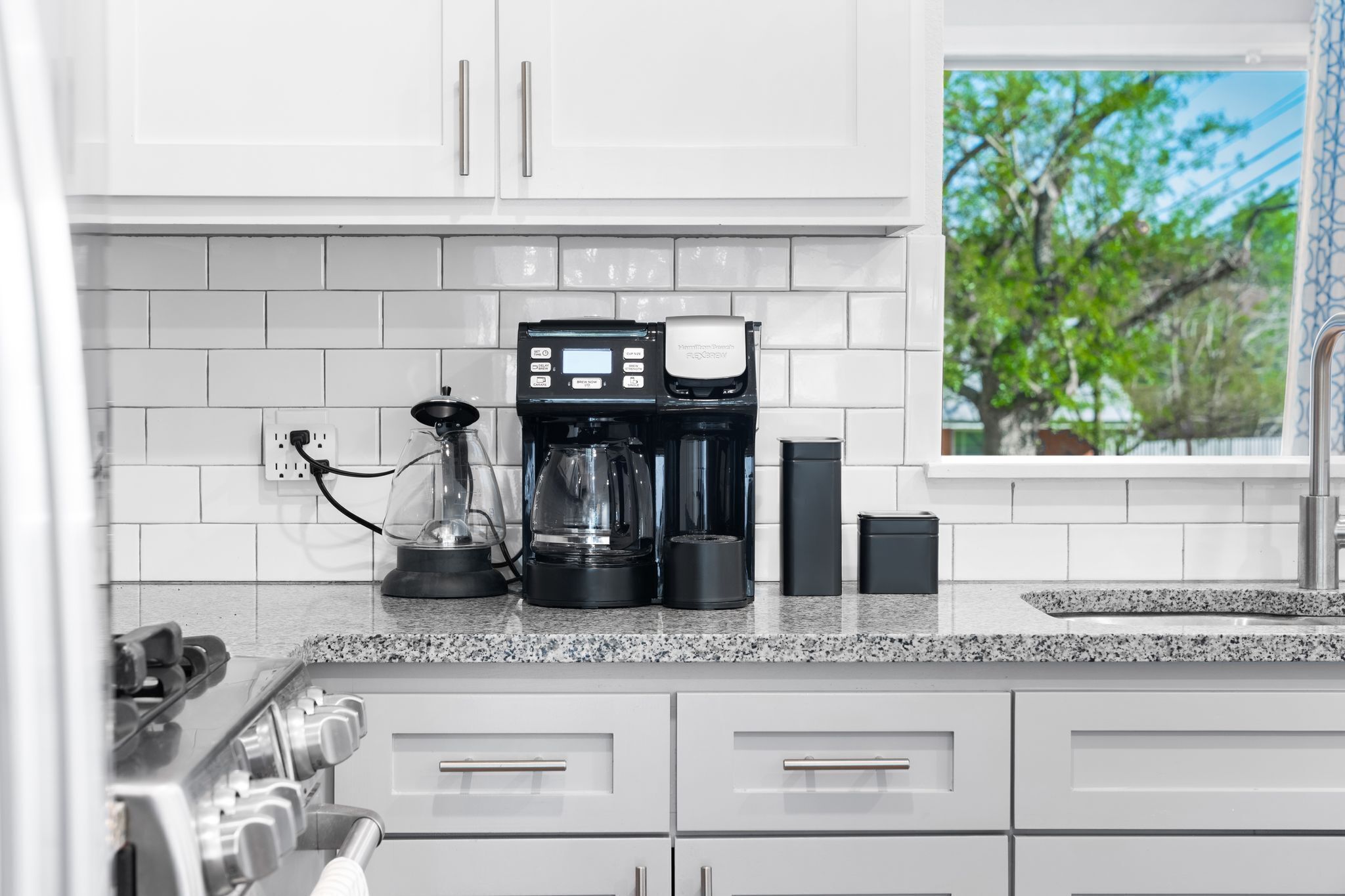 Une cuisine moderne avec des meubles blancs, un plan de travail en granit, une machine à café et une fenêtre donnant sur la forêt.