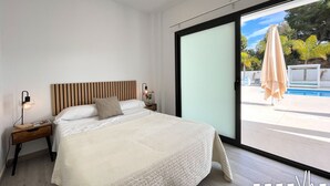 A bedroom with a large bed, a nightstand, a bedside lamp, and a view of a swimming pool through a sliding glass door. - MARTA BEACH 1 – Beachfront apartment in Benissa (Benissa)