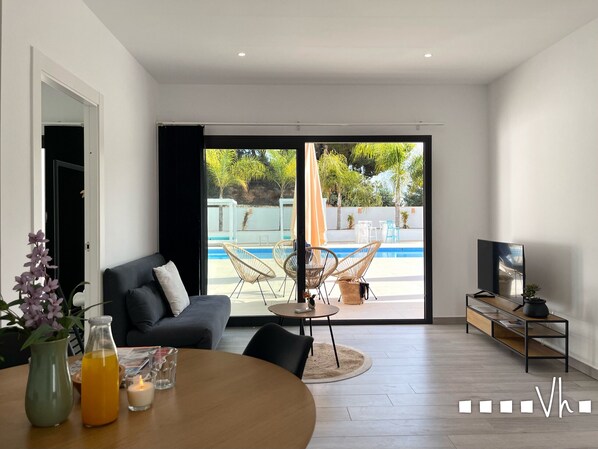 Un salon moderne avec un canapé, une table à manger avec des boissons, un téléviseur et une vue sur une piscine et du mobilier d’extérieur.