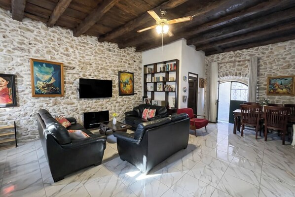 A stone-walled living room with a fireplace, a flat-screen TV, a bookshelf, and a dining area with wooden furniture.
