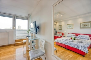 A bedroom with a bed, a desk, a chair, a TV, and a large window.