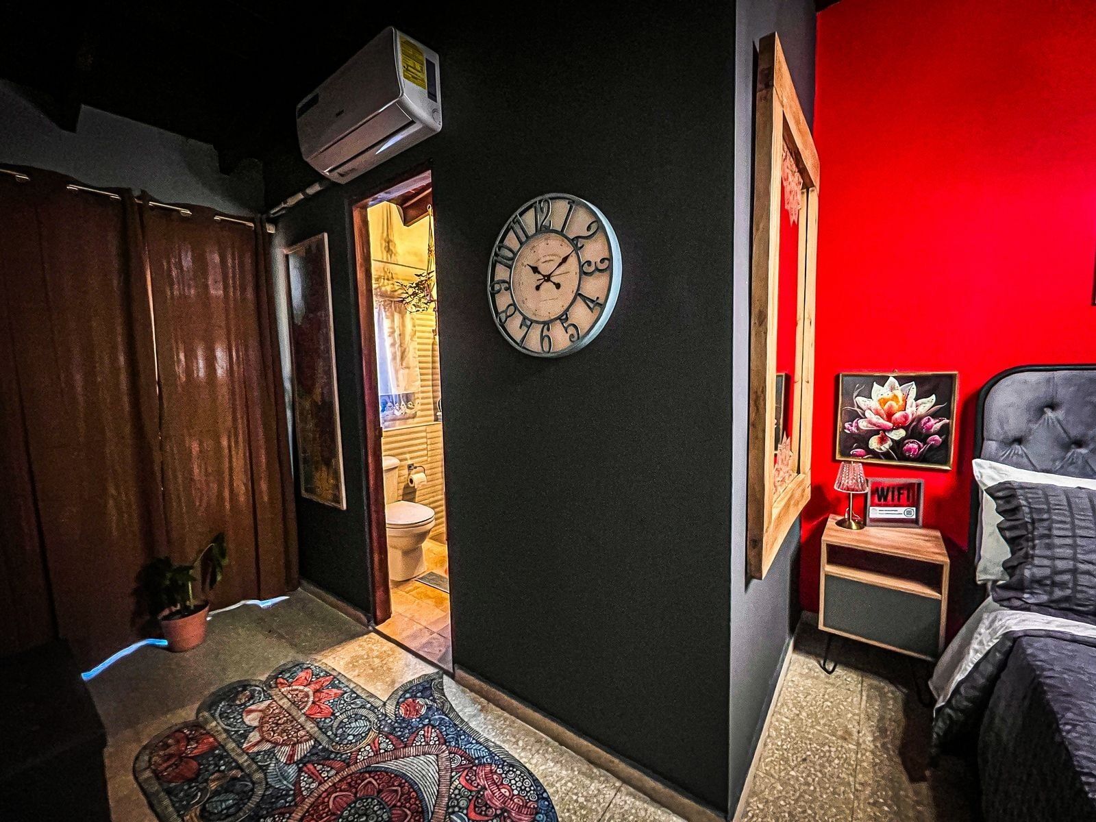 Un dormitorio con una cama, una mesita de noche con lámpara, un reloj y una pared roja.