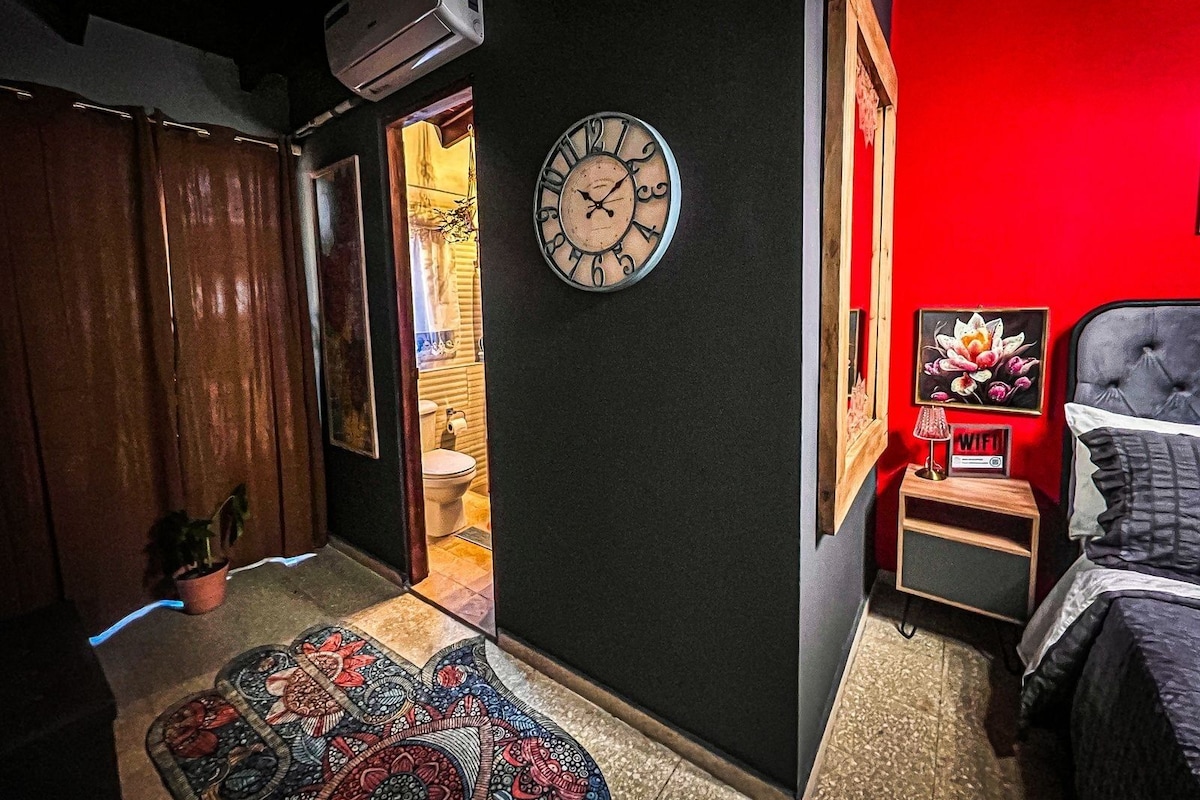 A bedroom with a bed, a bedside table with a lamp, a clock, and a red wall.
