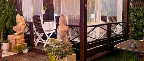 Um pátio externo com pérgola de madeira, luzes pendentes e duas estátuas.