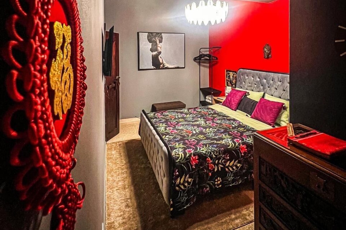 A bedroom with a bed, a red accent wall, a chandelier, and a decorative red wall hanging.