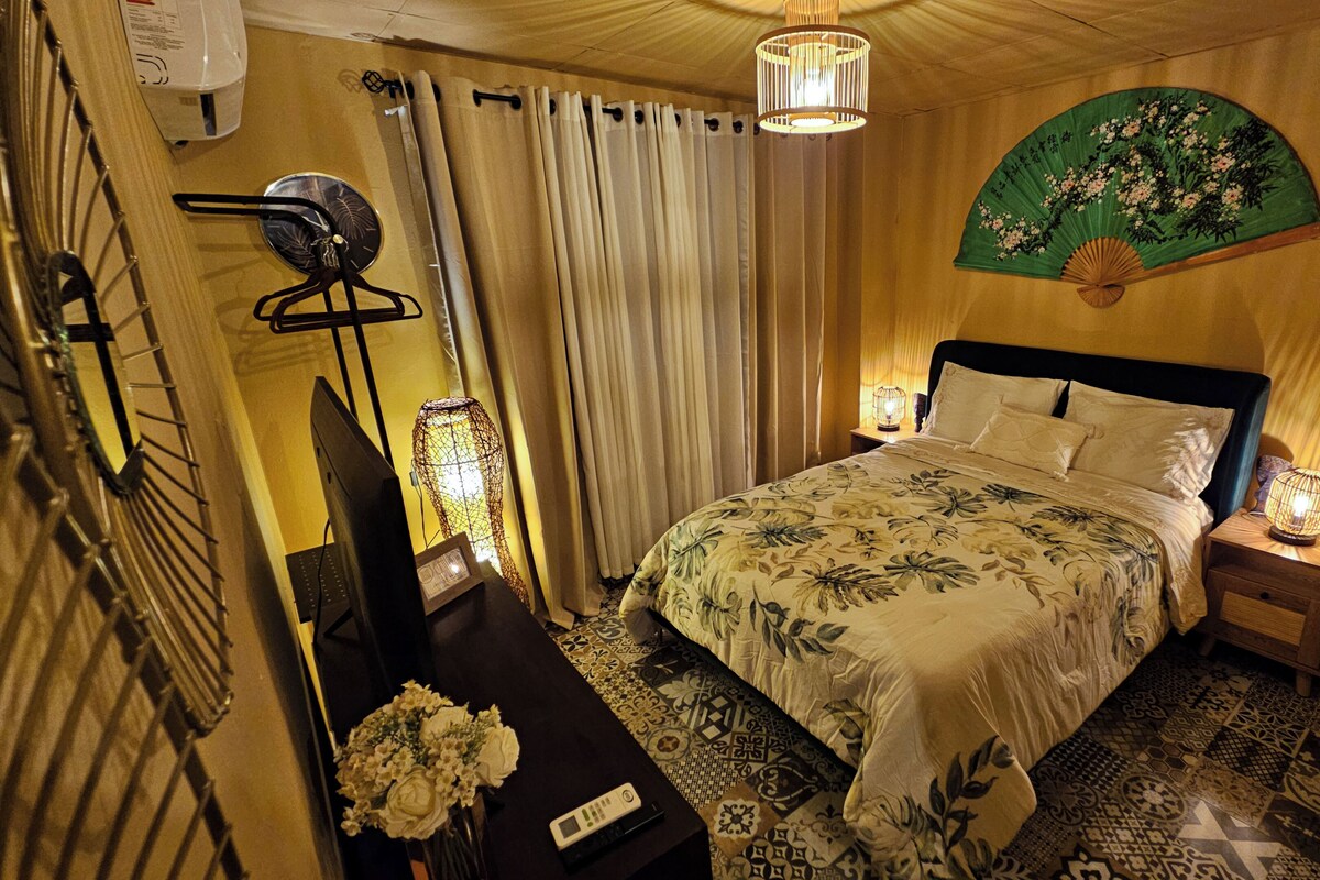 A hotel room with a bed, bedside lamps, a fan, a decorative fan, a wall-mounted clock, and a curtain.
