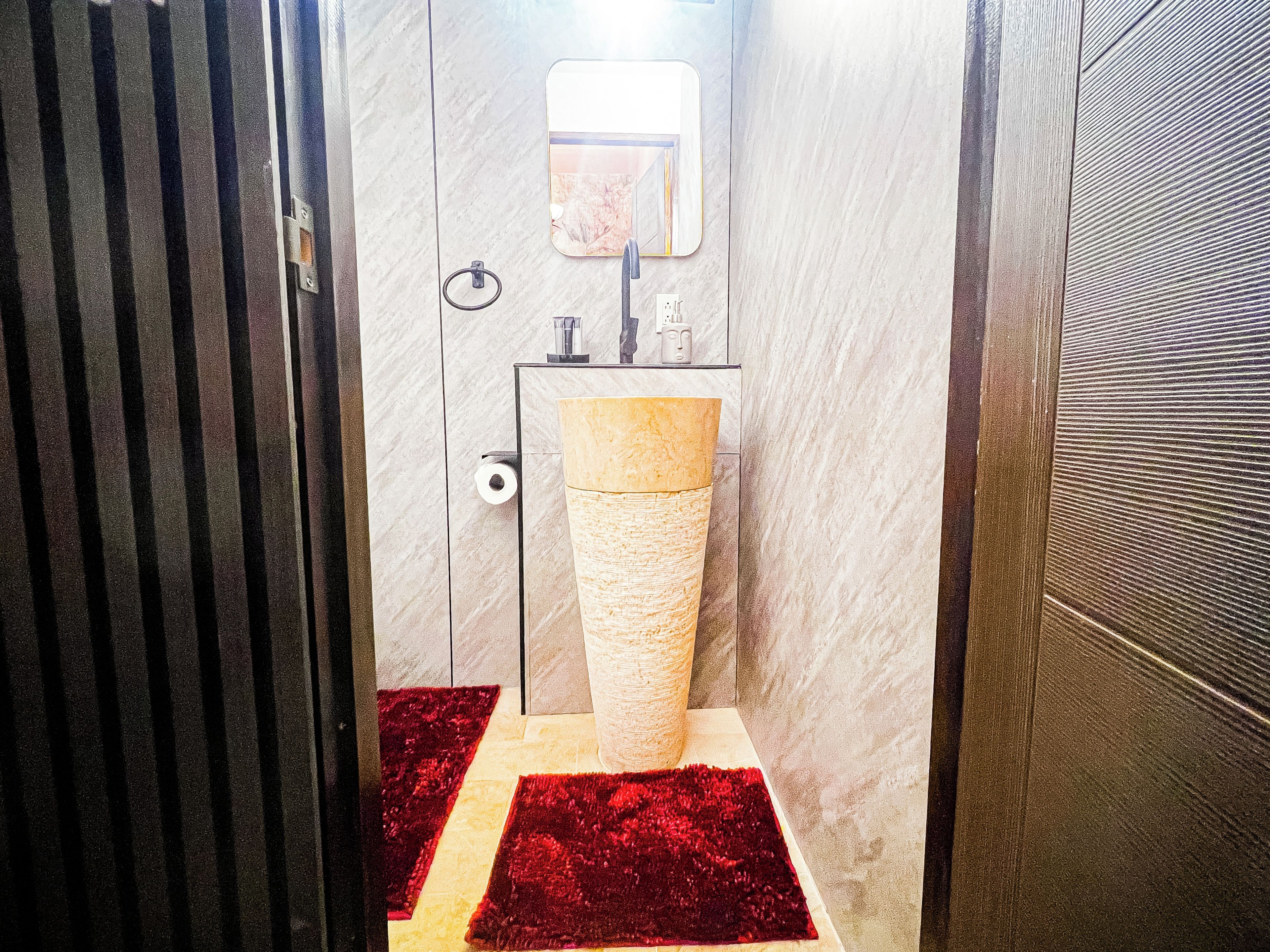 A bathroom with a marble wall, a sink with a beige basin, and a red rug on the floor.