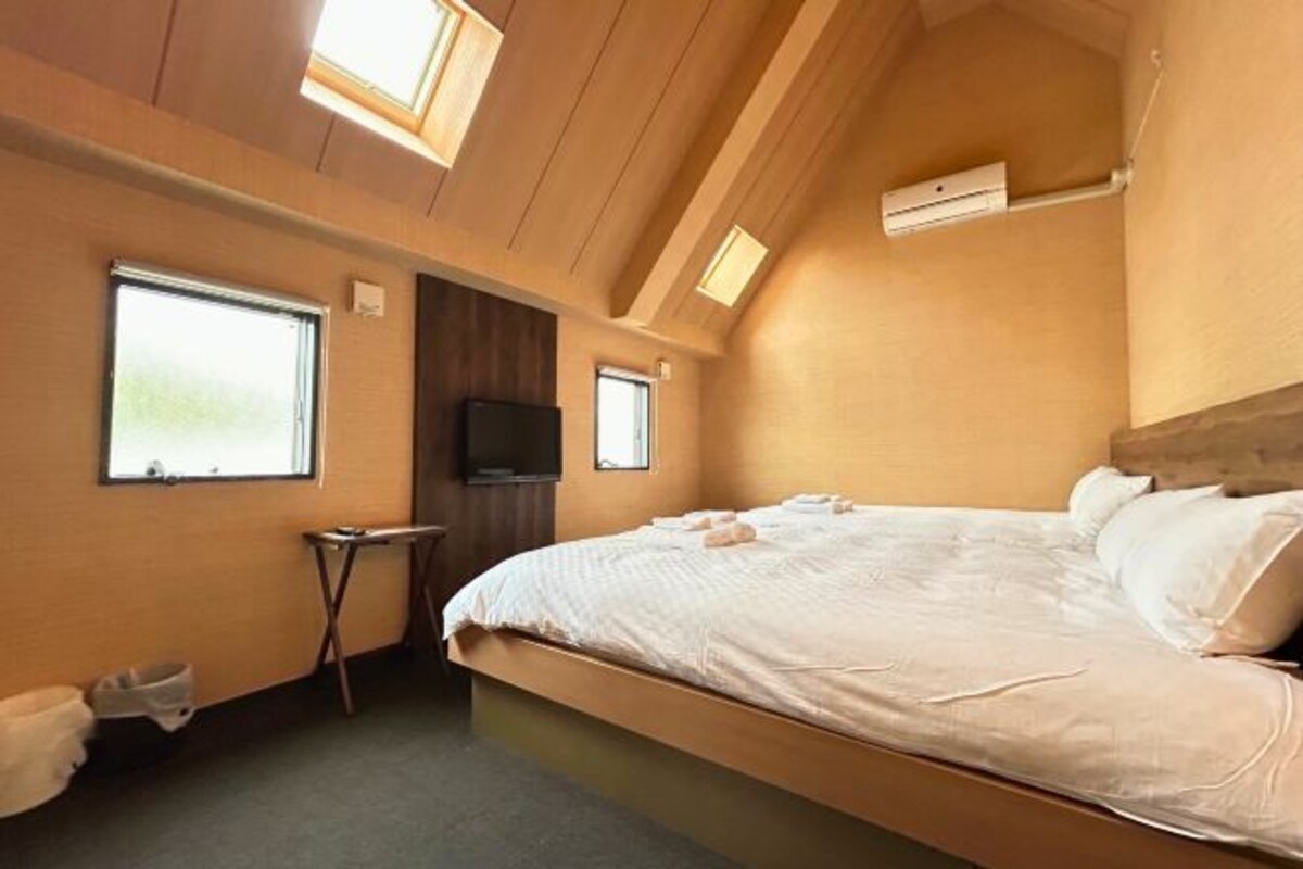 A bedroom with a large bed, two windows, a flat-screen TV, and a small table.