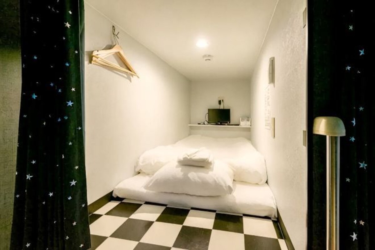 A room with a bed, a hanging coat rack, a small shelf with items, and a checkered floor.