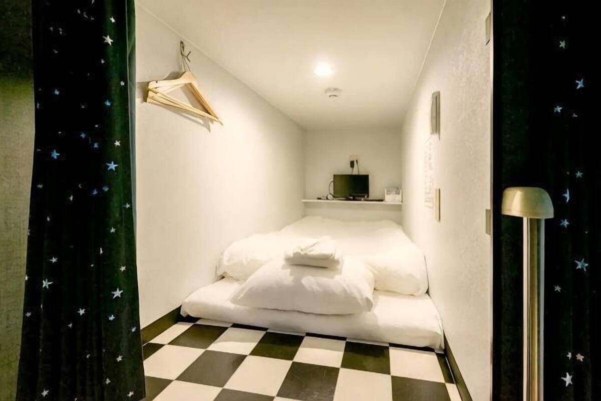 A room with a bed, a hanging coat rack, a small shelf with items, and a checkered floor.