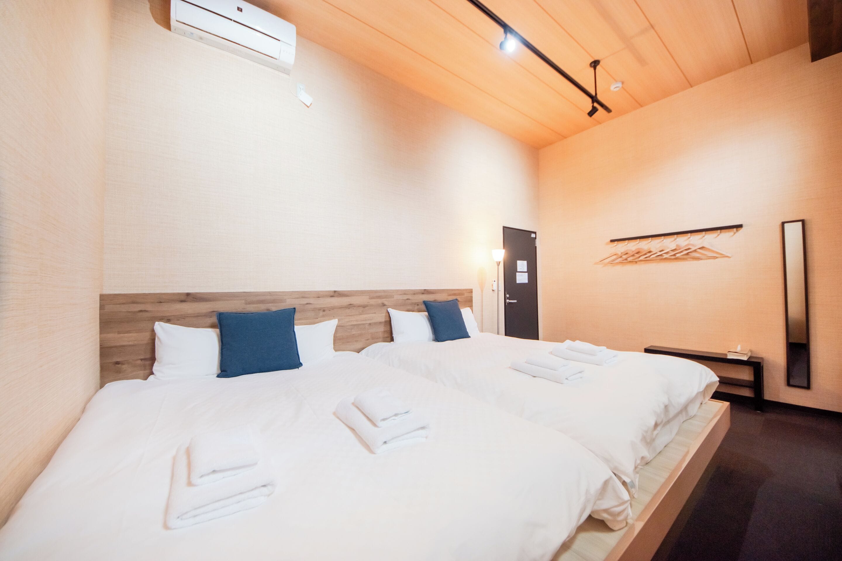 Quarto de hotel com duas camas, cabeceira de madeira, ar condicionado fixado na parede, mesa de cabeceira com abajur e um cabideiro.