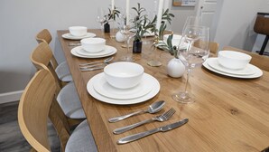 A dining table set with white plates, bowls, cutlery, and wine glasses, surrounded by wooden chairs with grey cushions.