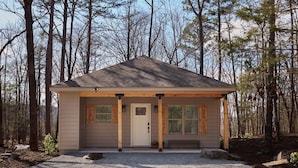 A small, single-story cabin with a gabled roof, a front porch with columns, and a gravel driveway.