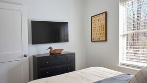 Een slaapkamer met een bed, een kast, een flatscreen televisie en een raam met zonwering.