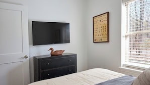 Ein Schlafzimmer mit Bett, Kommode, Flachbildfernseher und einem Fenster mit Jalousien.