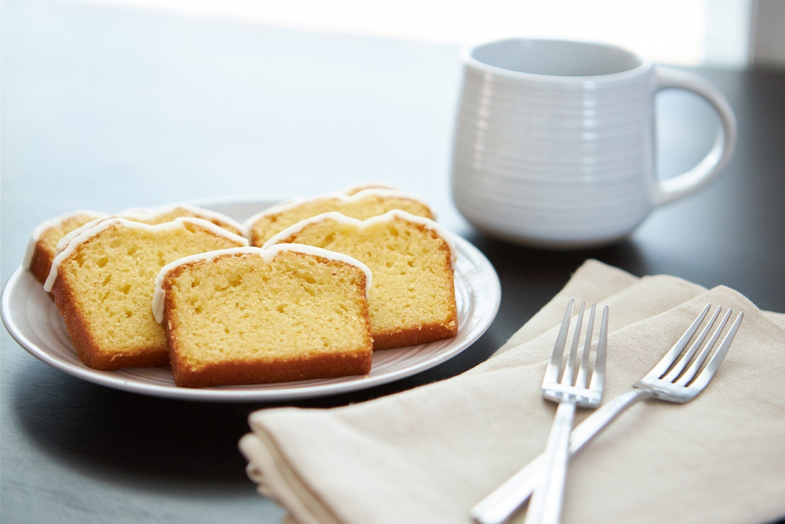 Een bord met plakjes in elkaar gezakt cake, een witte beker en een gevouwen servet met twee vorken op een donkere ondergrond.