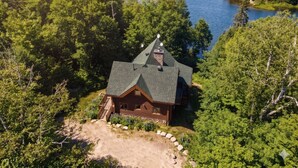 A cabin with a sloped roof, surrounded by dense forest and a lake.
