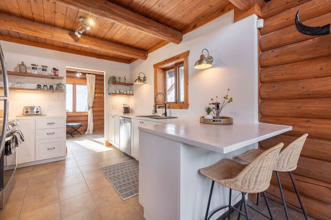 A kitchen with wooden beams, white cabinets, a central island, bar stools, and a window with curtains.
