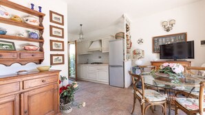 A kitchen with white cabinets, a refrigerator, a dining table with chairs, and a television. - VILLINO LA POLVERIERA (Porto Azzurro)
