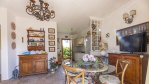 A dining area with a round glass table, floral chairs, a chandelier, and a television on the wall. - VILLINO LA POLVERIERA (Porto Azzurro)