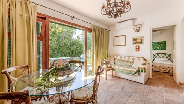 A dining area with a glass table and chairs, a sofa, and a bed in the adjacent room. - VILLINO LA POLVERIERA (Porto Azzurro)