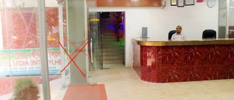 A hotel reception area with a marble counter, a receptionist, and a staircase leading to the upper floor.