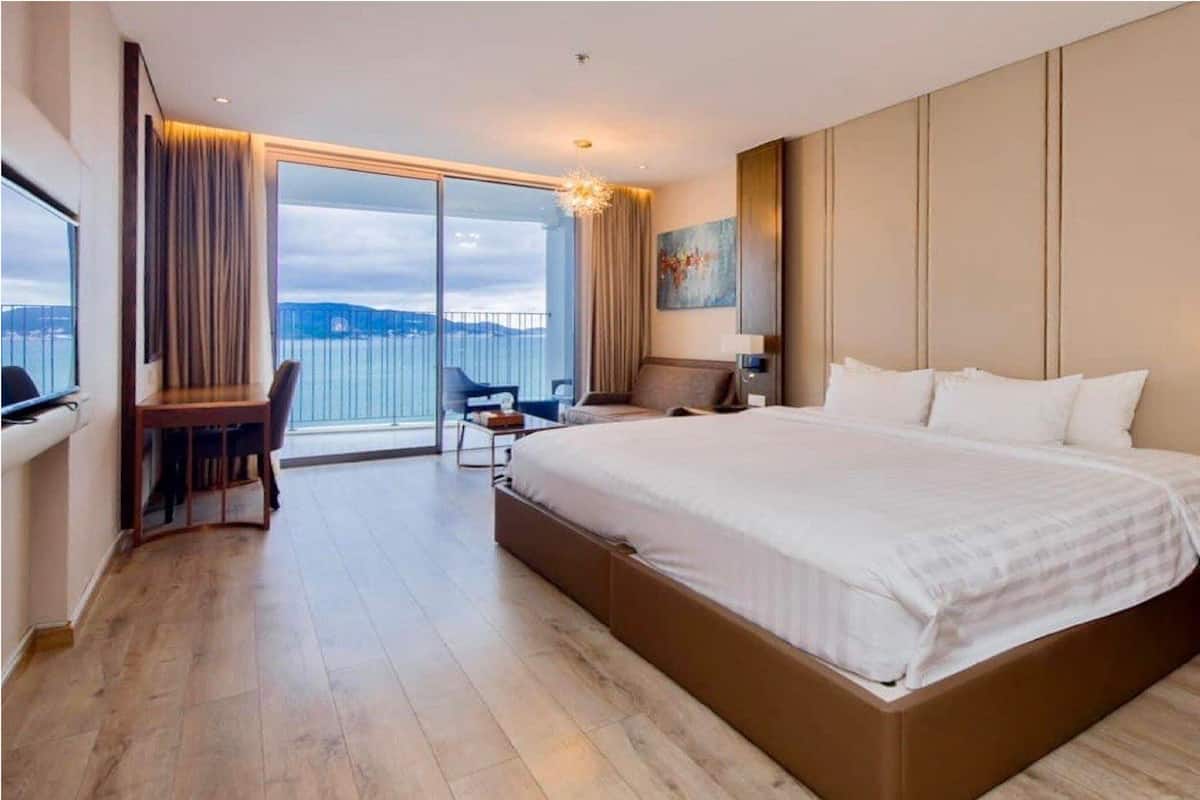 Une chambre d’hôtel moderne avec un grand lit, un bureau et une chaise. Une grande fenêtre offre une vue sur la mer.