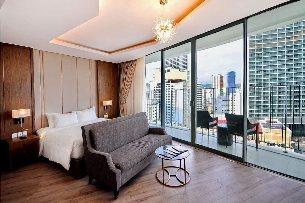 Une chambre d’hôtel moderne dotée d’un grand lit, d’un canapé, d’une petite table et offrant une vue sur la ville grâce à de grandes fenêtres du sol au plafond.