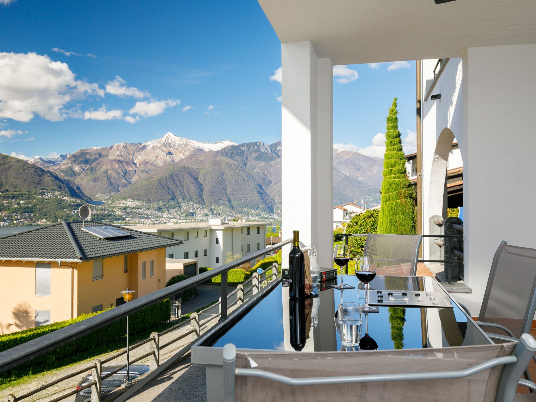 Ein Balkon mit Glastisch, Weinflasche und Gläsern, mit Blick auf eine Berglandschaft und Gebäude.